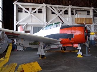 A vintage military aircraft is displayed inside a hangar. The airplane has a predominantly white body with distinctive red and black markings, featuring a red nose and a yellow propeller tip. The hangar shows structural beams and windows in the background, while a sign on the ground provides information about the aircraft.