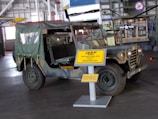 A vintage military Jeep from the Vietnam War era is on display in a museum. The Jeep has a camouflage pattern and an open-top design with a canvas covering at the back. It is parked indoors, and a large yellow sign in front of it provides details about the Jeep. The museum setting includes other historical exhibits and aircraft, visible in the background.