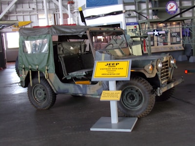 A vintage military Jeep from the Vietnam War era is on display in a museum. The Jeep has a camouflage pattern and an open-top design with a canvas covering at the back. It is parked indoors, and a large yellow sign in front of it provides details about the Jeep. The museum setting includes other historical exhibits and aircraft, visible in the background.