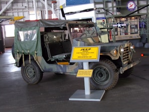 A vintage military Jeep from the Vietnam War era is on display in a museum. The Jeep has a camouflage pattern and an open-top design with a canvas covering at the back. It is parked indoors, and a large yellow sign in front of it provides details about the Jeep. The museum setting includes other historical exhibits and aircraft, visible in the background.