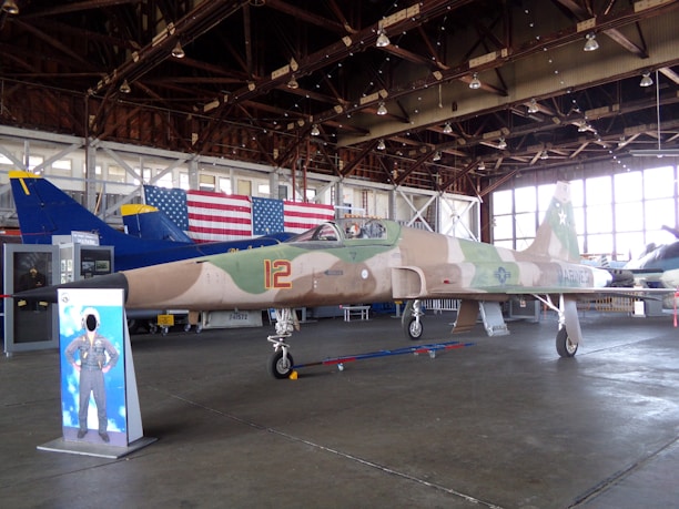 A camouflaged military jet with the number 12 on its side is displayed inside a large hangar. Behind the jet is a large American flag and other aircraft with blue and yellow tails. The hangar has a high ceiling with exposed beams and hanging lights. There is a cut-out standee of a pilot in front of the jet, and some informative displays can be seen near the aircraft.