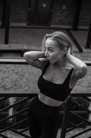 A woman in a black athletic outfit rests her arms on a railing, with her head tilted slightly and hands touching her hair. The setting features a brick walkway and a wooden bench behind her, with a focus on her contemplative expression and graceful posture.