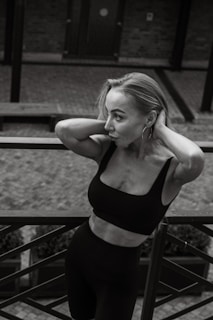 A woman in a black athletic outfit rests her arms on a railing, with her head tilted slightly and hands touching her hair. The setting features a brick walkway and a wooden bench behind her, with a focus on her contemplative expression and graceful posture.