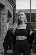 A woman standing outdoors against a brick and glass building background, wearing a dark jacket over a black sports bra and leggings. She has long hair and is looking slightly upwards with a confident expression.