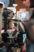 A person is preparing coffee using a pour-over method. They are holding a gooseneck kettle and pouring hot water into a dripper over a glass carafe. The setting appears to be in a coffee shop with an espresso machine nearby.