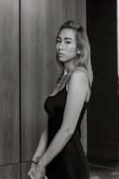 A woman wearing a minimalist black dress paired with sleek accessories posing against a cream-colored wall.