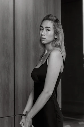 A woman wearing a minimalist black dress paired with sleek accessories posing against a cream-colored wall.