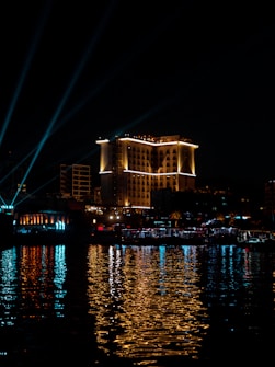 A well-lit hotel building stands prominently at night. Surrounding it are smaller structures with neon and warm lights reflecting beautifully on the body of water in the foreground. The night sky is dark, contrasting with the bright architectural lighting and reflections.