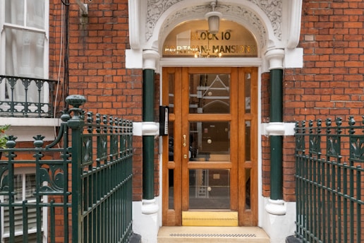 A brick building entrance with an arched top, featuring a wooden door with glass panels and surrounded by decorative iron fencing. The words 'Block 10 Portman Mansions' are displayed above the door.