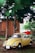 A vintage yellow Volkswagen Beetle car is parked among tall trees with a wooden house in the background. A red jeep is partially visible behind the Beetle. The setting appears rustic, with elements of greenery and natural surroundings.