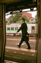 A man in a suit is walking briskly on a train platform, holding a briefcase. In the background, a commuter train is visible, with greenery and urban buildings.