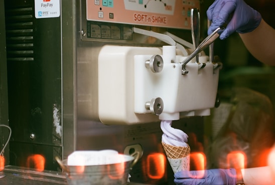 A person wearing blue gloves operates a soft serve ice cream machine, with the ice cream being dispensed into a waffle cone. The background includes some machine controls and an indicator of a payment system.