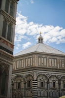 A historic baptistery with intricate geometric patterns and detailed arches is seen against a bright blue sky with scattered clouds. The architecture features ornate details and a symmetrical design with a distinct dome and decorative facade.