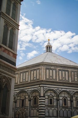 A historic baptistery with intricate geometric patterns and detailed arches is seen against a bright blue sky with scattered clouds. The architecture features ornate details and a symmetrical design with a distinct dome and decorative facade.