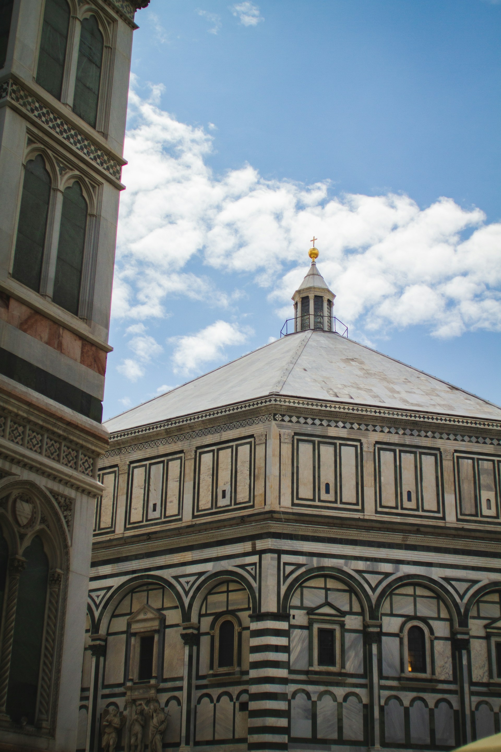 A historic baptistery with intricate geometric patterns and detailed arches is seen against a bright blue sky with scattered clouds. The architecture features ornate details and a symmetrical design with a distinct dome and decorative facade.
