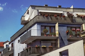 An apartment building featuring multiple balconies with railings, each adorned with potted plants and flowers. The building has a modern design with blue and gray tones, and a satellite dish is visible on one of the top balconies. A bright blue sky with a few clouds serves as the backdrop.