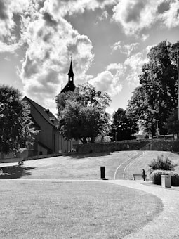 A peaceful park setting features a large grassy area with a winding path, lined with trees and a bench. In the background, a church with a tall spire is silhouetted against a partly cloudy sky. A lone figure walks up the path, adding a sense of tranquility to the scene.