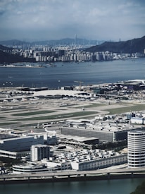 An aerial view of an urban landscape featuring a large airport with multiple runways and terminals. In the foreground, buildings and industrial structures are visible, while in the background, a vast body of water separates the airport from a densely packed city with many skyscrapers and a mountainous backdrop.
