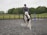 A person riding a white horse with brown patches in an outdoor equestrian arena. The rider is wearing a helmet and casual riding attire. The arena is enclosed by a wooden fence and has several jumping poles placed in the background. The setting appears to be a large, grassy field with trees in the background under a cloudy sky.
