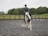 A person riding a white horse with brown patches in an outdoor equestrian arena. The rider is wearing a helmet and casual riding attire. The arena is enclosed by a wooden fence and has several jumping poles placed in the background. The setting appears to be a large, grassy field with trees in the background under a cloudy sky.
