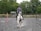 A person dressed in equestrian gear, including a helmet, is riding a white and brown horse on a gravel surface in an outdoor setting. The background features lush green trees and a fenced area, indicative of an equestrian training ground or arena.