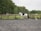 A person is riding a horse in an outdoor riding arena surrounded by fencing. The horse is white with brown patches. The rider is wearing equestrian gear, including a helmet. In the background, there is green foliage and trees.