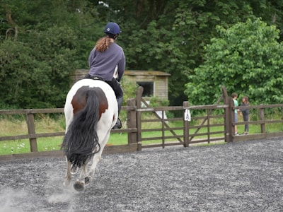 A person is riding a horse on a gravel surface inside a fenced area. The rider is wearing a helmet and casual riding clothes. The horse is primarily white with dark patches. In the background, two people are standing by a wooden gate, surrounded by lush green trees and foliage.