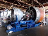 A large jet engine is displayed on a blue support structure inside a museum or exhibition hall. Many wires and components are visible on the engine, suggesting detailed engineering. In the background, panels with various posters and images are arranged on the wall.