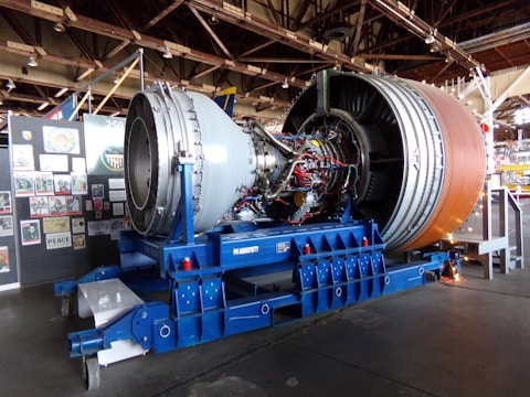 A large jet engine is displayed on a blue support structure inside a museum or exhibition hall. Many wires and components are visible on the engine, suggesting detailed engineering. In the background, panels with various posters and images are arranged on the wall.