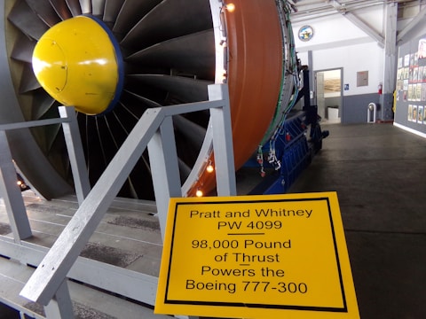 A large jet engine on display in an indoor setting, with a prominent yellow and blue cone at the center of the engine. The engine is surrounded by a gray wooden platform with railings, and a yellow informational sign in the foreground describes the engine's specifications. The background shows a hallway lined with various posters or images on the walls.