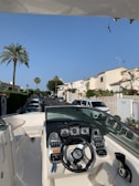 A boat is parked in a residential street, showing its dashboard with multiple gauges and a steering wheel. The street is lined with white houses, and cars are parked along the road. Palm trees and greenery add to the scenery, under a clear blue sky.