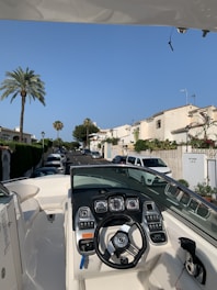 A boat is parked in a residential street, showing its dashboard with multiple gauges and a steering wheel. The street is lined with white houses, and cars are parked along the road. Palm trees and greenery add to the scenery, under a clear blue sky.