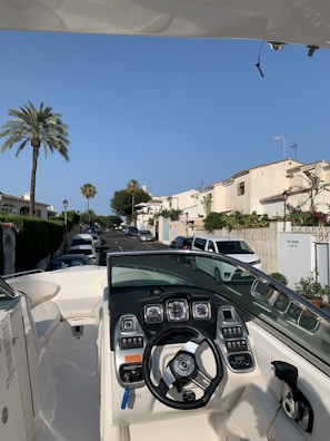 A boat is parked in a residential street, showing its dashboard with multiple gauges and a steering wheel. The street is lined with white houses, and cars are parked along the road. Palm trees and greenery add to the scenery, under a clear blue sky.