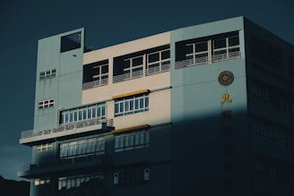 A multi-story building with a large facade featuring windows and balconies is partially illuminated by sunlight, casting dramatic shadows across the structure. The building exterior is painted in a light blue and gray color scheme. On the facade, there is a circular emblem and some Chinese characters in yellow.