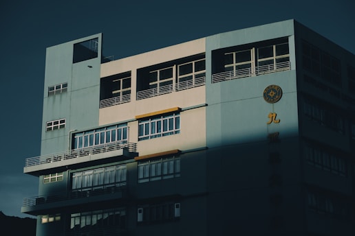 A multi-story building with a large facade featuring windows and balconies is partially illuminated by sunlight, casting dramatic shadows across the structure. The building exterior is painted in a light blue and gray color scheme. On the facade, there is a circular emblem and some Chinese characters in yellow.