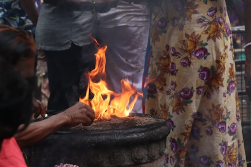 A fire burning brightly in an outdoor setting, surrounded by a group of people. One person's hand is seen tending to the fire, and colorful clothing with floral patterns can be observed.