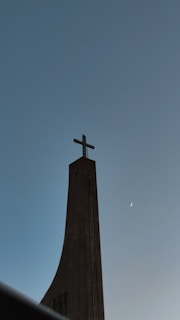 A tall, modern church steeple with a prominent cross at the top, set against a clear evening sky with a visible crescent moon to the right.
