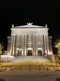 A grand neoclassical building with a row of tall columns across the front, illuminated at night. There is a central staircase leading up to three large, ornate doors. Above the columns, a Latin inscription reads 'SOLI DEO HONOR ET GLORIA' with statues positioned at the top.