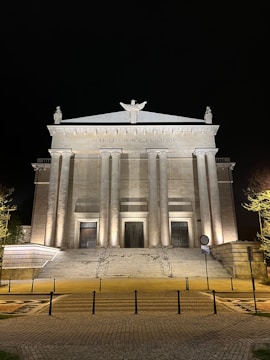 A grand neoclassical building with a row of tall columns across the front, illuminated at night. There is a central staircase leading up to three large, ornate doors. Above the columns, a Latin inscription reads 'SOLI DEO HONOR ET GLORIA' with statues positioned at the top.