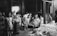 A group of workers, primarily young men, are gathered around piles of fibrous material, possibly jute or hemp, in a dimly lit industrial setting. The scene suggests manual labor, with some individuals directly engaging with the material while others stand to the side. The space appears large and warehouse-like, indicating a factory environment.