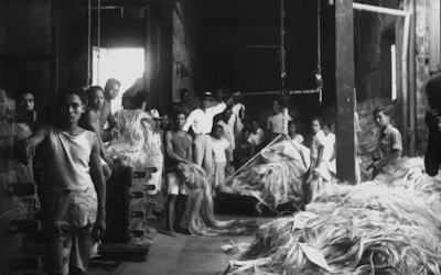 A group of workers, primarily young men, are gathered around piles of fibrous material, possibly jute or hemp, in a dimly lit industrial setting. The scene suggests manual labor, with some individuals directly engaging with the material while others stand to the side. The space appears large and warehouse-like, indicating a factory environment.