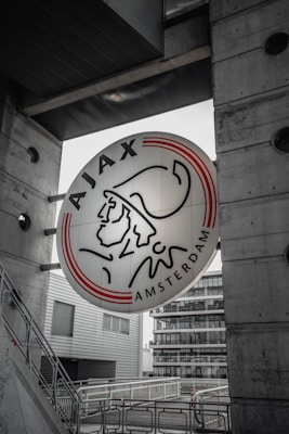 A large round sign featuring a stylized illustration of a historical figure wearing a headpiece is mounted between two concrete structures. The text 'AJAX AMSTERDAM' encircles the illustration. In the background, there are modern buildings and geometric architecture with glass and metal elements.