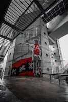 A large mural on a concrete wall depicts a figure wearing a number 14 jersey, standing and pointing. The mural features a red and black color scheme with distinctive white crosses. The surroundings are industrial with metallic structures and geometric designs, creating an urban atmosphere.