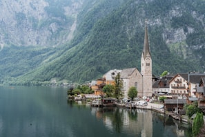 A picturesque lakeside village nestled against steep, lush green mountains, with a prominent clock tower and spire rising from a church amid quaint houses. The calm water reflects the natural beauty and the buildings, creating a serene and tranquil atmosphere.