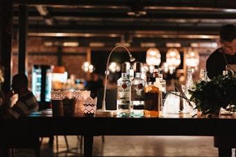 A bar counter in a dimly lit setting displaying several bottles of liquor, including Bacardi, alongside cocktail-making tools. Decorative lanterns adorn the counter, creating a cozy ambiance. In the background, large hanging lights provide additional warm lighting, and a bartender is partially visible.