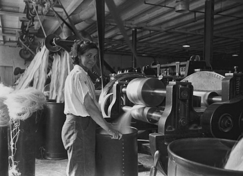 A woman is operating machinery in a textile factory. She stands next to a large machine with rollers and levers, working with fiber materials. The factory has high ceilings and other machinery is visible in the background.