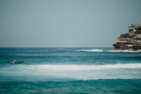 The image presents a serene seascape with several people surfing on waves. On the right side, there is a rocky cliff partially covered with green vegetation. The sky is clear, and the ocean has varying shades of blue and turquoise.
