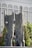 A large outdoor stone sculpture features two tall, abstract rock forms with smooth surfaces and cut-out holes. Figures of people are integrated into the sculpture, standing or seated. The backdrop includes a modern building with glass windows and columns, and greenery like trees.