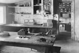 A vintage workshop with wooden paneling features a large workbench covered with various tools and materials. Shelves on the walls hold numerous boxes and supplies, while a hanging lamp provides lighting. The scene conveys a sense of organized clutter typical of a busy workspace.