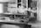 A vintage workshop with wooden paneling features a large workbench covered with various tools and materials. Shelves on the walls hold numerous boxes and supplies, while a hanging lamp provides lighting. The scene conveys a sense of organized clutter typical of a busy workspace.